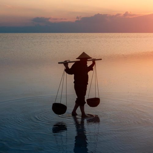 person carrying water buckets in rural lake setting during sunset